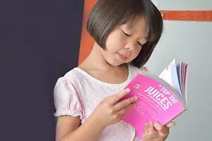 A young girl with short hair wearing a white polka-dot dress is holding a book titled 'The Top 100 Juices'.