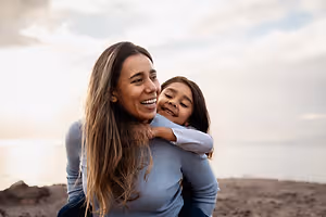 A woman with long hair holds a smiling girl in her arms on a beach under a cloudy sky