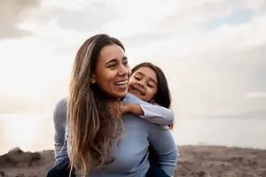 A woman with long hair holds a smiling girl in her arms on a beach under a cloudy sky