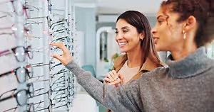 Two women are smiling and looking at a display of eyeglasses in a store.