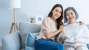 A woman and an elderly woman are sitting on a couch. The woman is smiling and holding a cup. The elderly woman is smiling and hugging the woman. Behind them is a lamp, a picture frame, and a plant in a vase. The lamp is on the left, and the plant is on the right.