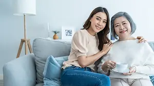 A woman and an elderly woman are sitting on a couch. The woman is smiling and holding a cup. The elderly woman is smiling and hugging the woman. Behind them is a lamp, a picture frame, and a plant in a vase. The lamp is on the left, and the plant is on the right.