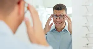 A man is adjusting his glasses in a mirror, smiling and looking at himself.