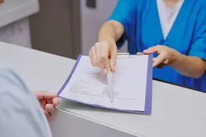 A person in blue scrubs hands a paper with a pen to another person in a light blue shirt.