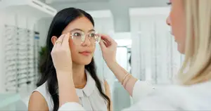 A woman is trying on glasses in a store with the assistance of another woman wearing a white shirt and a bracelet