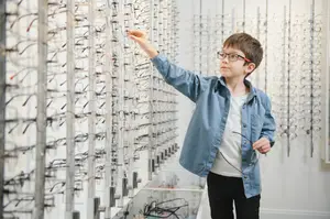 A young boy with glasses is looking at different glasses on a wall in a store.