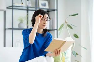 A woman wearing glasses is sitting on a couch and reading a book while adjusting her glasses with her left hand.
