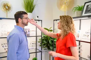 A man is adjusting his glasses in a room with a woman looking at him.