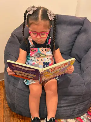 A little girl with glasses sits on a beanbag chair reading a book