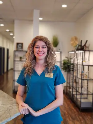 A smiling woman in blue scrubs stands inside an office with wooden floors and shelves