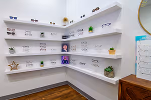 A corner of an eyeglass shop with a display of glasses on white shelves and potted plants