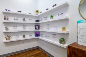 A corner of an eyeglass shop with a display of glasses on white shelves and potted plants