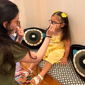 A woman is adjusting a young girl's glasses in a room with patterned cushions.