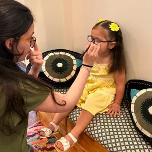 A woman is adjusting a young girl's glasses in a room with patterned cushions.