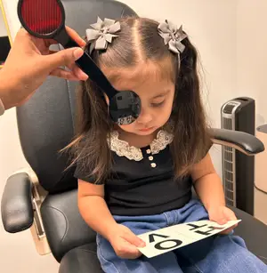 A young girl is sitting on a chair holding a reading chart with letters and is being examined by a person using a magnifying tool.