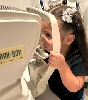 A young girl with a bow in her hair is looking through a white ophthalmoscope with the text Dark-800 on it.