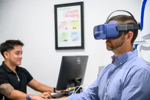 A man wearing a blue virtual reality headset and looking down, and another man is sitting in front of a computer monitor.