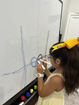 A little girl in a yellow dress and glasses draws on a whiteboard with blue markers.