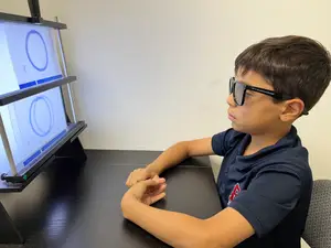 A young boy wearing glasses and sitting in front of a desk with a computer monitor displaying a circle image