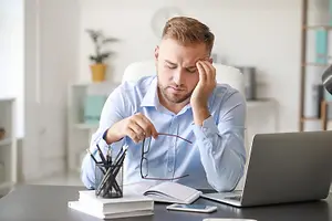A man is sitting on a chair and holding his head with his right hand, probably in the office. He is looking at something on the desk, which has a book, a laptop, a cell phone, and a pen holder with pens. Behind him, there is a shelf with a plant and a lamp, and on the right side, there is a desk with a lamp. In the distance, there is a white wall.
