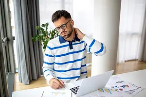 A man sitting at a desk with a laptop and papers, rubbing his neck