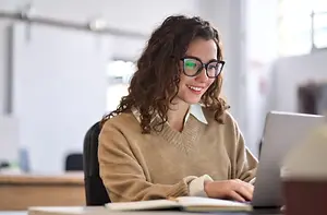 A woman with curly hair wearing glasses is sitting on a chair in front of a desk with a laptop and an open book, smiling and possibly posing for a photo in an office with a whiteboard and glass window in the background.
