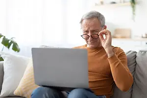 An old man is sitting on a couch, wearing glasses, and holding a laptop on his lap while touching his glasses.