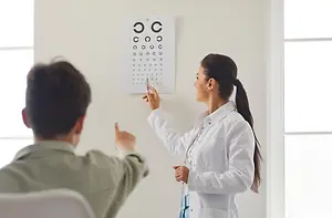 A woman in a white lab coat demonstrates an eye chart to a man in a green shirt.