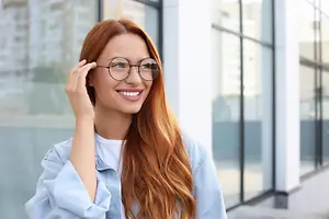 A woman with red hair and glasses is smiling and posing for a photo outside a building.