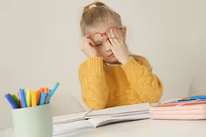 Tired young girl rubbing her eyes while wearing glasses at a desk with school supplies