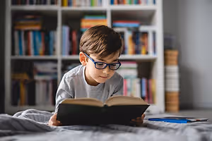Child wearing glasses reading a book on a bed