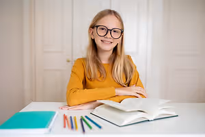 Smiling girl wearing glasses at a desk with an open book and colored pencils