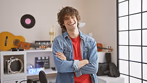A smiling man in a denim jacket and red shirt is standing with his arms crossed in a room filled with musical instruments, including a guitar, a trumpet, a violin, and a drum set, and a black vinyl record is hanging on the wall. The room has a white shelf with a drum set, a guitar, a trumpet, and a violin on it. A neon sign that reads 'ON AIR' is on the left side of the shelf. The room has a glass window with a black frame. 