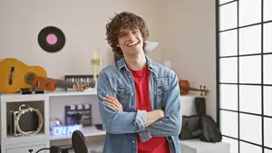 A smiling man in a denim jacket and red shirt is standing with his arms crossed in a room filled with musical instruments, including a guitar, a trumpet, a violin, and a drum set, and a black vinyl record is hanging on the wall. The room has a white shelf with a drum set, a guitar, a trumpet, and a violin on it. A neon sign that reads 'ON AIR' is on the left side of the shelf. The room has a glass window with a black frame. 