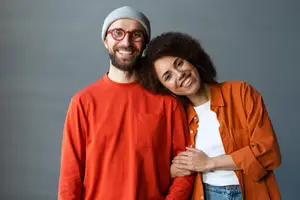 A man and a woman wearing orange clothes are standing next to each other, smiling, and posing for a photograph in front of a gray wall.