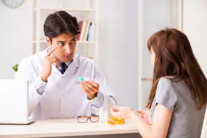 A man in a lab coat showing eye drops to a woman in a clinic.