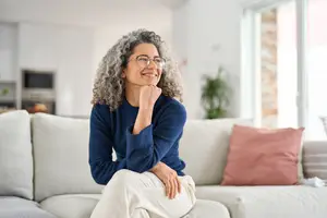 A woman with curly gray hair wearing glasses and a blue sweater is sitting on a couch with a smile on her face.