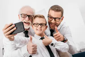 Three people posing for a photo, including an older man holding a phone, a boy in a white shirt, and a man in glasses with a watch on his wrist.