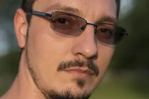 A man with a mustache and beard wearing glasses with a serious expression.