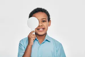 A young boy wearing a blue polo shirt is holding a mirror and covering his left eye with a white circle, possibly posing for a photo.
