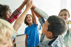 A group of children are in a classroom, raising their hands in the air while smiling.