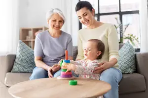An older woman and a young woman are sitting on a couch with a baby in front of them, playing with a toy.