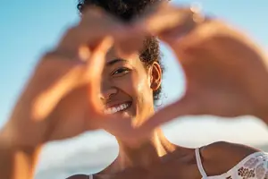 A woman wearing a white lace bra smiles and poses for a photo while making a heart shape with her hands
