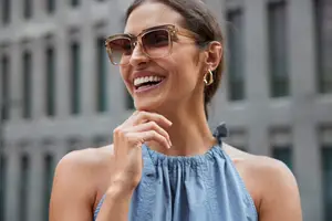A woman in sunglasses smiles with her hand on her chin in front of a building.