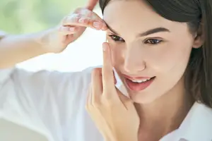 A woman with brown hair wearing a white shirt is applying makeup to her eyes while smiling and looking down. She is holding her eyebrows with her right hand and her left hand is on her cheek.