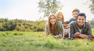 A family is lying on the grass in a park with a dog and smiling at the camera