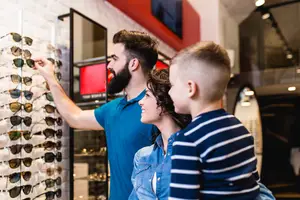 A family of three standing in front of a sunglasses rack in a store, the man is pointing at the sunglasses