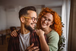 A man and a woman are smiling and posing for a photo in their home. The man has a beard and is wearing glasses. The woman has curly hair and is wearing a green sweater. Behind them is a white curtain and a plant. On the left side, there is a wooden table and chairs. On the right side, there is a window with a white curtain.