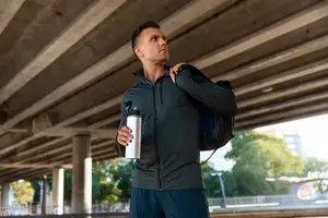 A man in a green jacket and blue shorts is standing under a bridge and holding a water bottle.