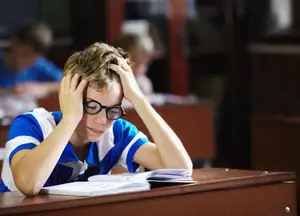 A boy with glasses in a classroom appears to be having a headache.